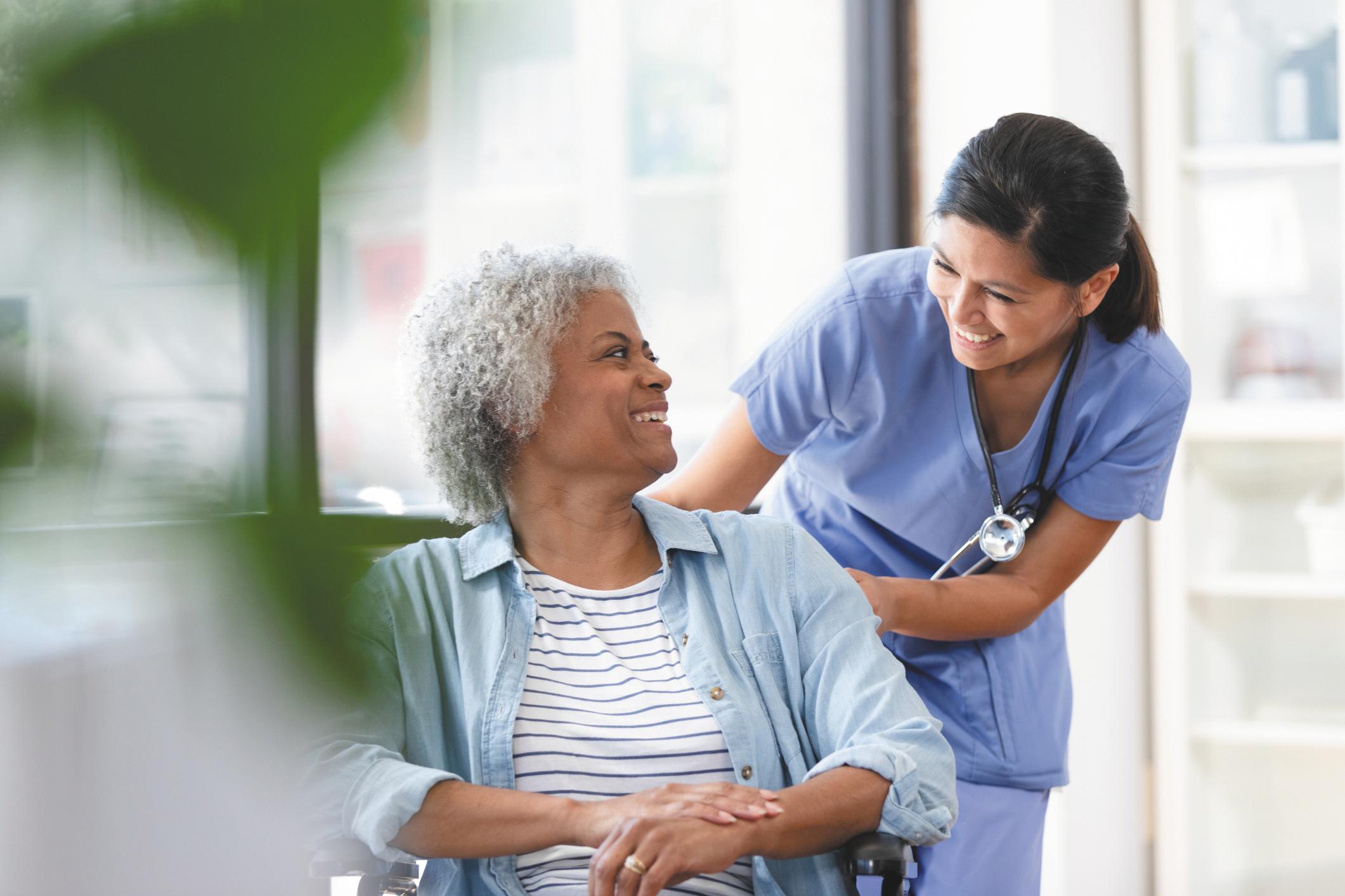 A nurse smiles while pushing a woman's wheelchair into an examination room.