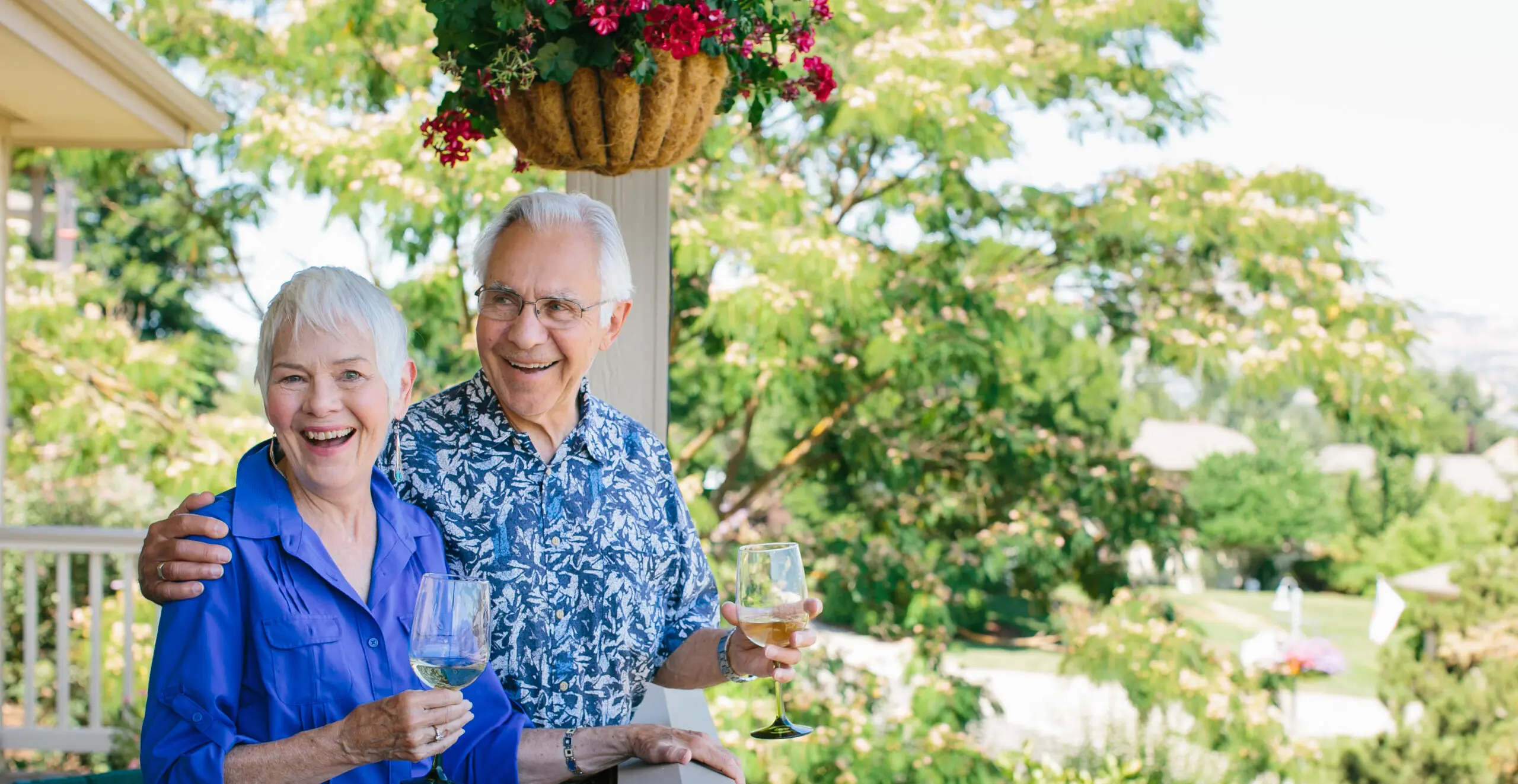 An older couple holding wine glasses smiles on an outdoor patio.
